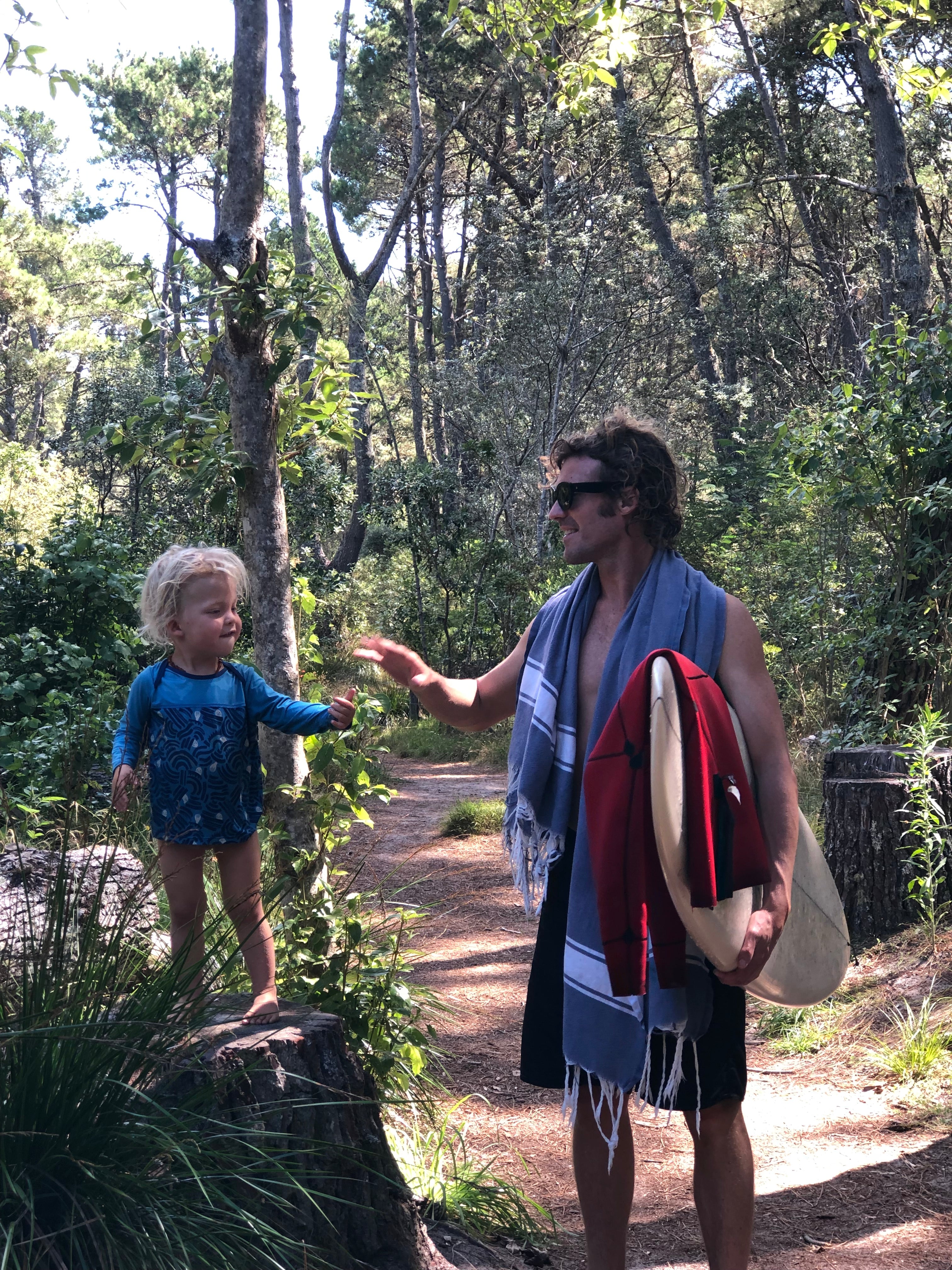 Man holding a surfboard standing amongst trees with a child