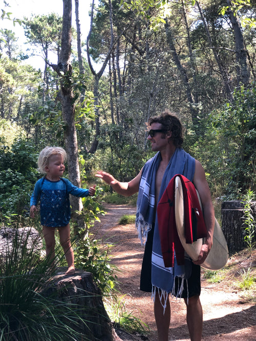 Man holding a surfboard standing amongst trees with a child